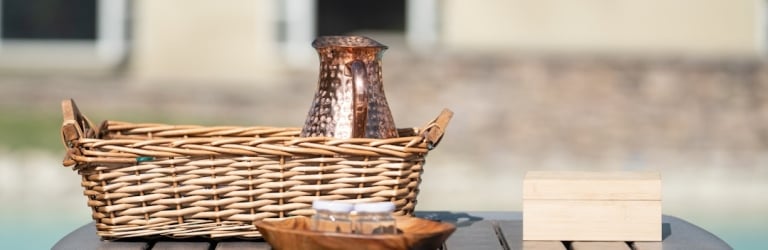 A wicker basket holding a copper pitcher, a wooden bowl, and a wooden box rest on a slatted outdoor table by a pool, a building blurred behind.