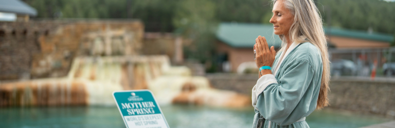 A woman with long gray hair, eyes closed, stands with hands pressed together in a light blue robe by a blue hot spring. Text: SOAKING MOTHER SPRING WORLD'S DEEPEST HOT SPRING DAILY GRATITUDE CEREMONY ALL ARE WELCOME.