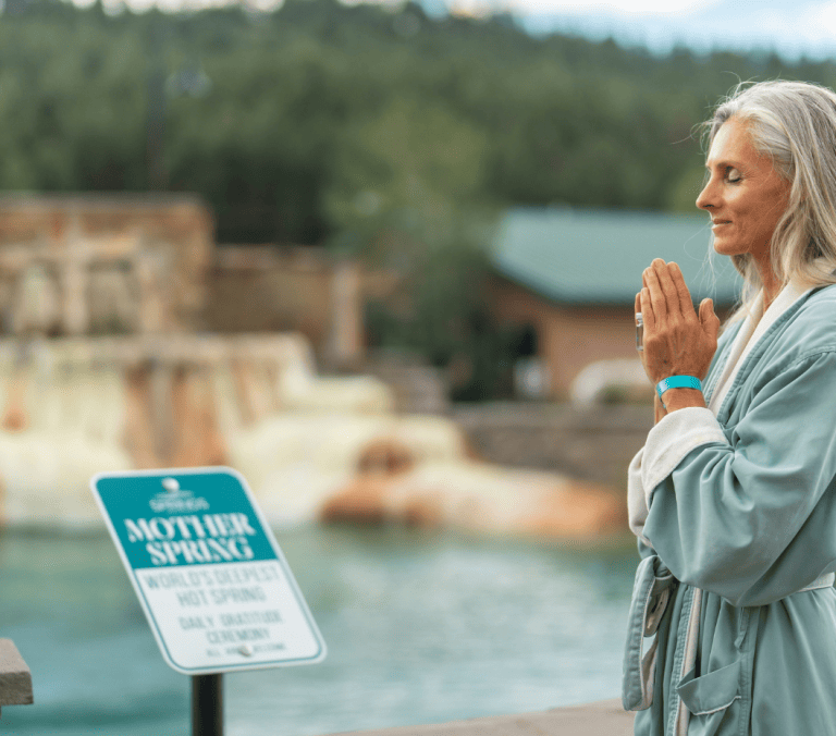 A woman with long gray hair, eyes closed, stands with hands pressed together in a light blue robe by a blue hot spring. Text: SOAKING MOTHER SPRING WORLD'S DEEPEST HOT SPRING DAILY GRATITUDE CEREMONY ALL ARE WELCOME.