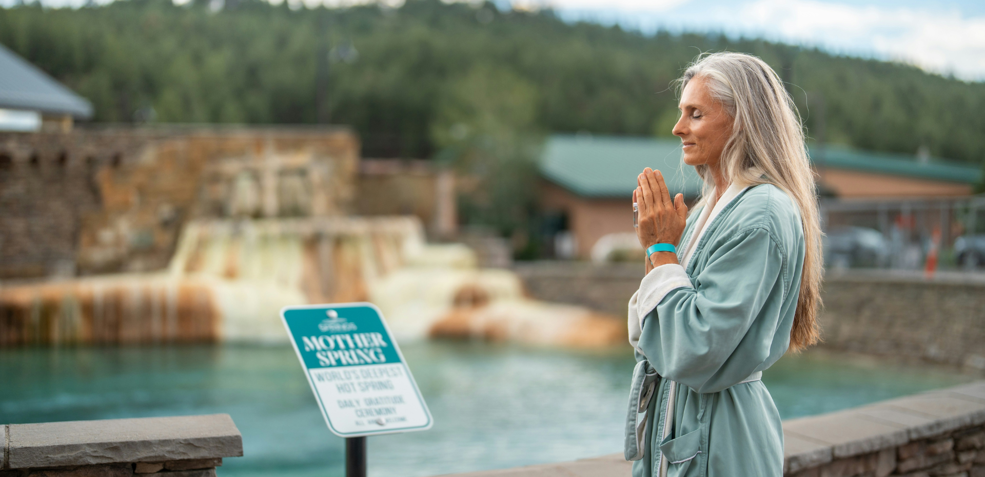 A woman with long gray hair, eyes closed, stands with hands pressed together in a light blue robe by a blue hot spring. Text: SOAKING MOTHER SPRING WORLD'S DEEPEST HOT SPRING DAILY GRATITUDE CEREMONY ALL ARE WELCOME.
