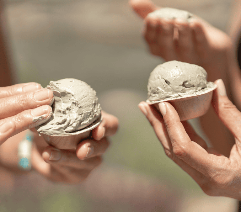 Hands scoop and hold light gray mud from small wooden bowls, with other hands and mud visible in the background, outdoors.