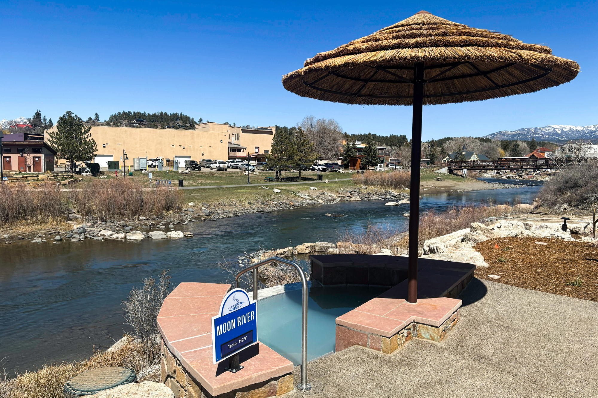 A hot tub with a large straw umbrella is positioned beside a river, with distant buildings and snow-capped mountains under a clear sky. Sign reads: MOON RIVER, Temp: 112°F.