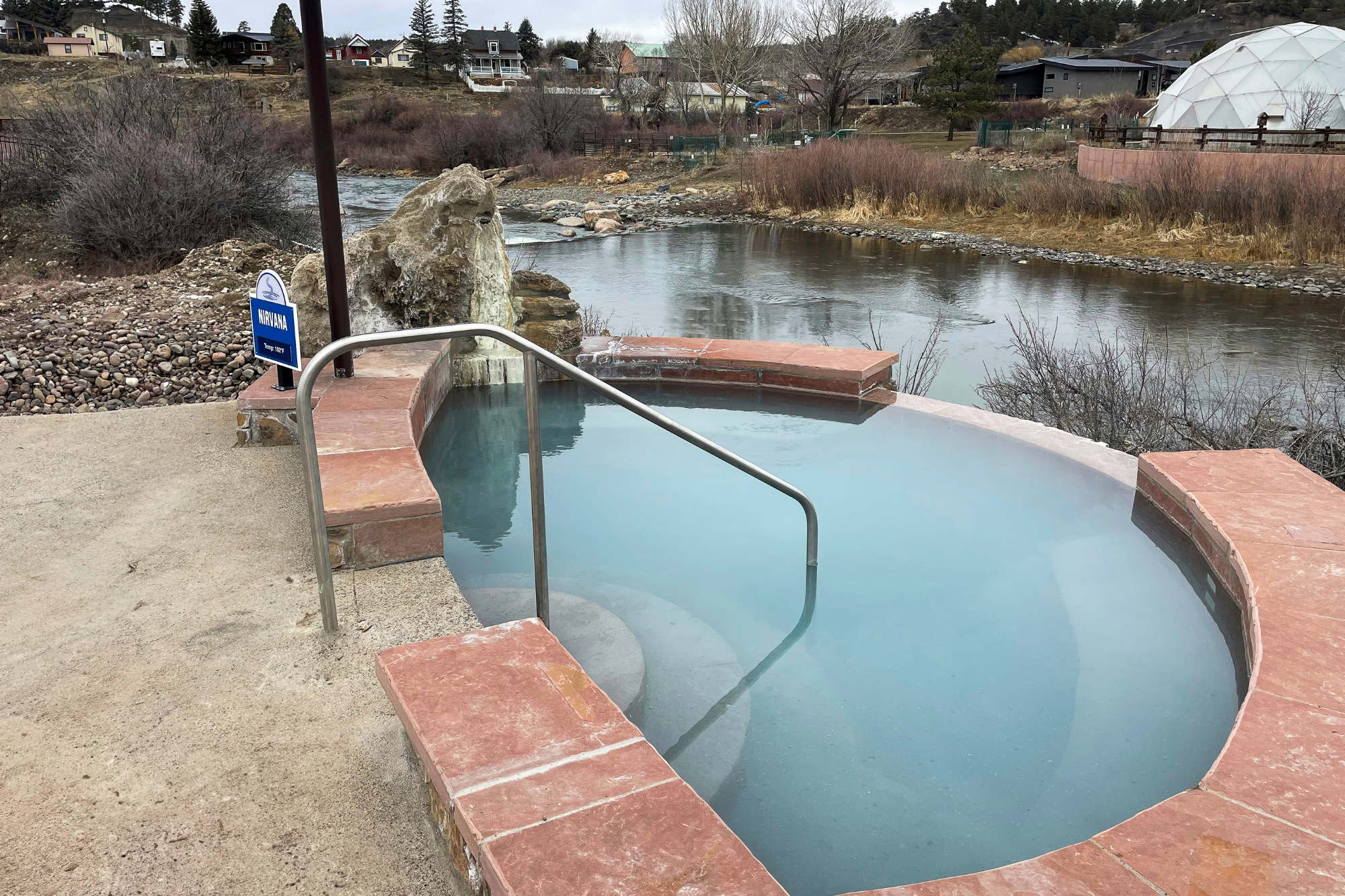 A light blue hot spring pool with red brick coping and a metal handrail overlooks a river, with distant houses on a rocky, bare hillside. Sign: NIRVANA Temp: 108°F.