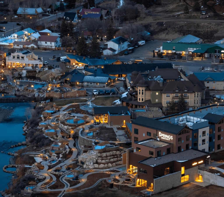 A large hot springs resort complex, featuring numerous illuminated pools and buildings, stretches along a riverbank amidst a town at dusk. Text: THE SPRINGS RESORT, GEOTHERMAL HEATING.