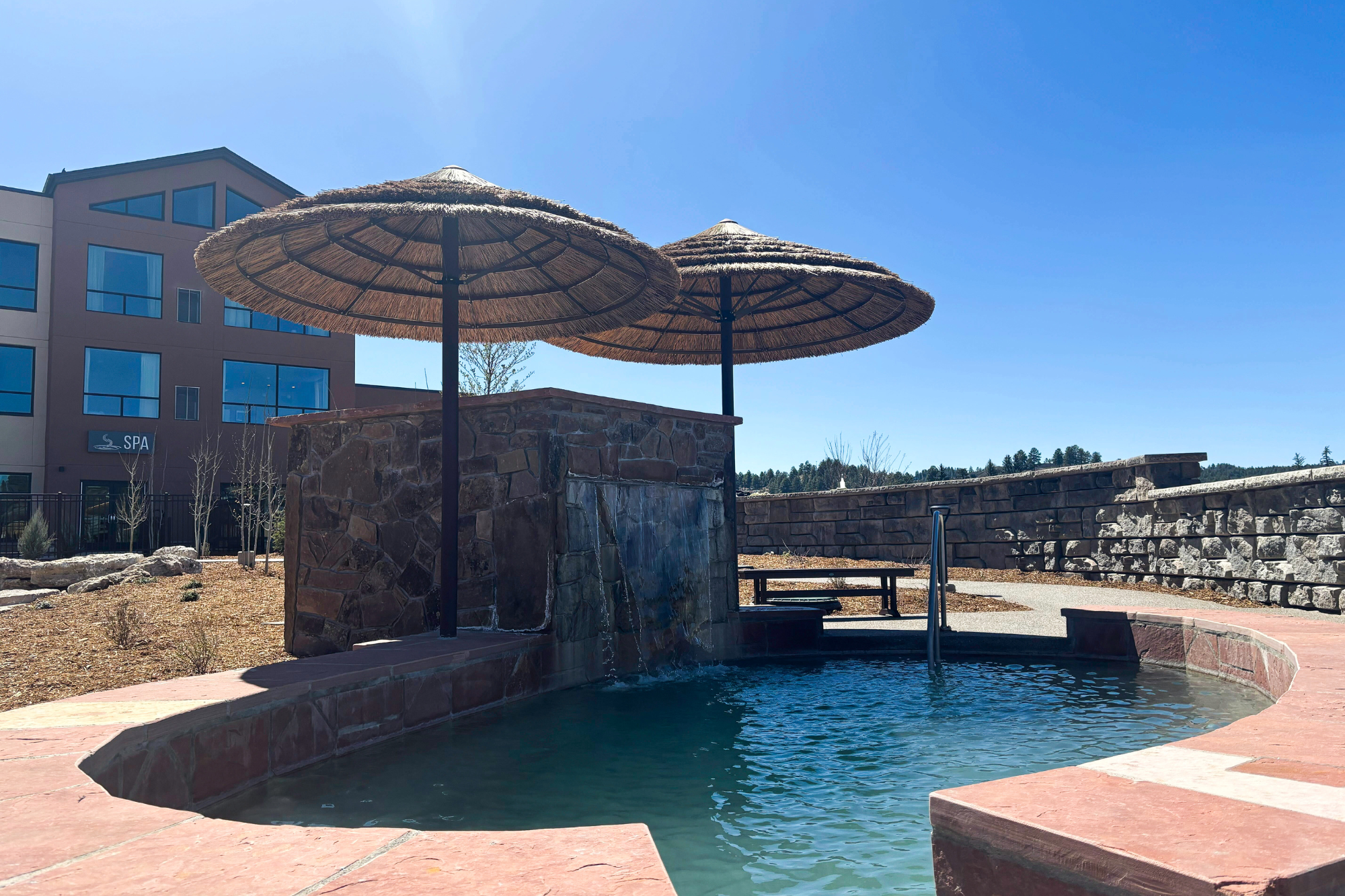 A curved hot tub with two straw umbrellas has water flowing from a stone waterfall. A modern building with a 