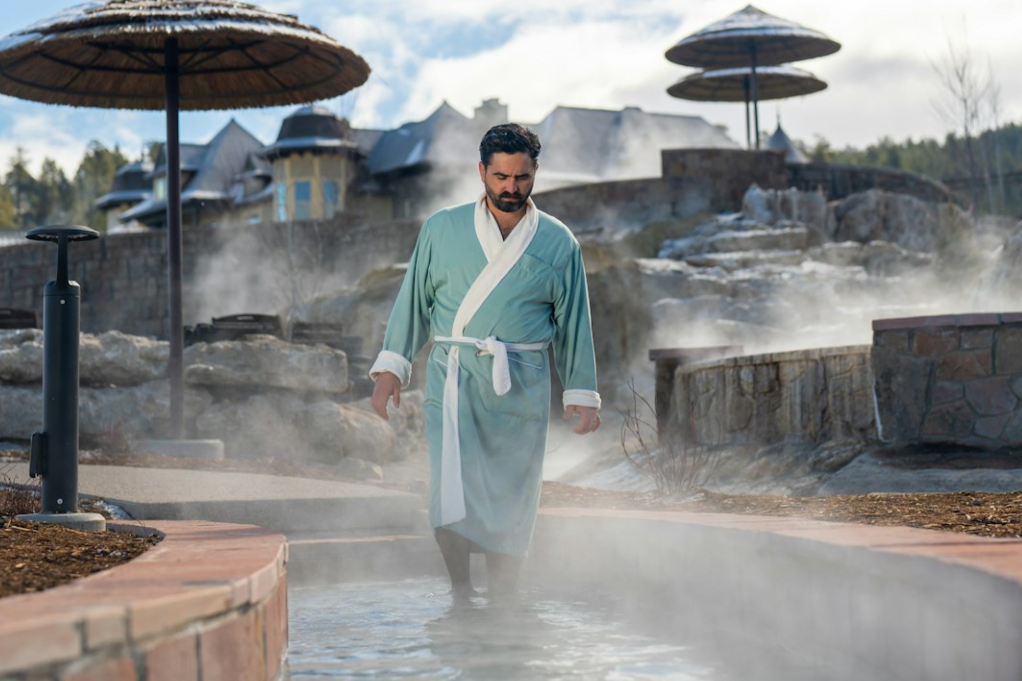 A man walks through a steaming outdoor pool, wearing a teal and white robe, surrounded by stone features and thatched umbrellas under a cloudy sky.
