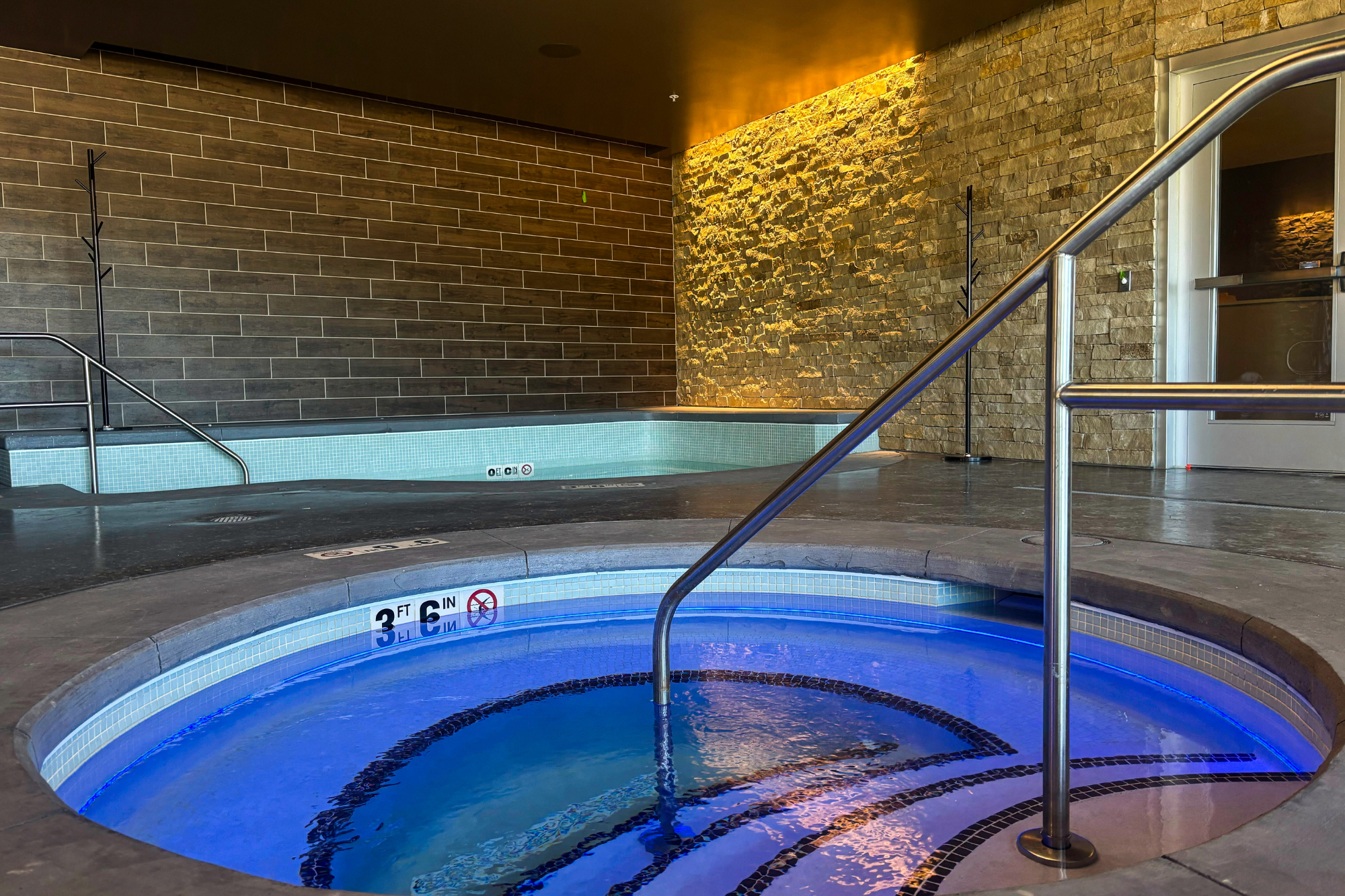 A round hot tub with blue water and a handrail displays '3 FT 6 IN', no diving, and no running symbols. A second rectangular hot tub, showing '0 FT C IN' and the same warnings, sits behind it in a room with dark tiled and stone walls.