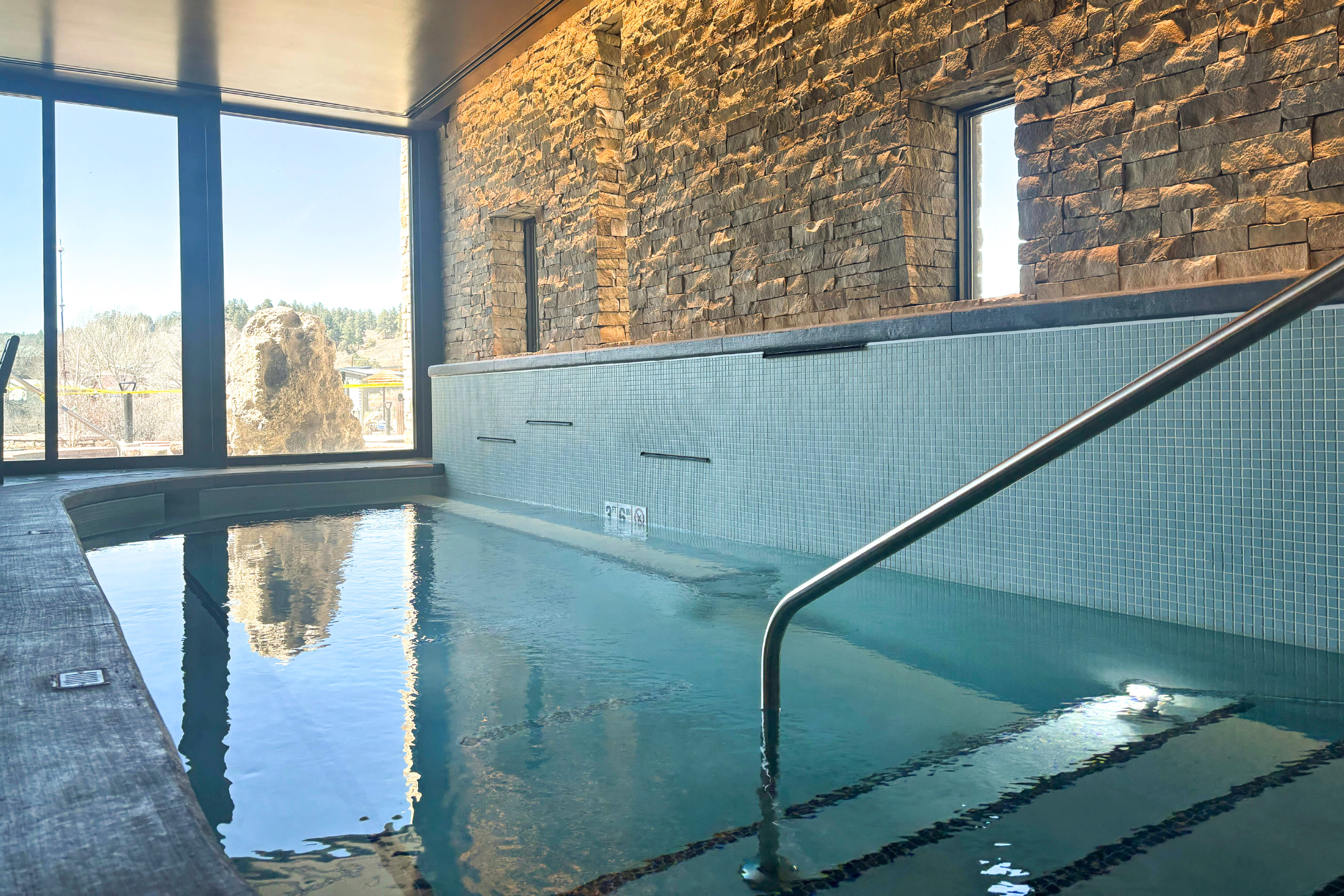 An indoor pool with tranquil blue water and a metal handrail, reflecting a sunny outdoor landscape through large windows. Stone and tiled walls surround it. Text: 3 FT 6 IN, No Diving symbol.