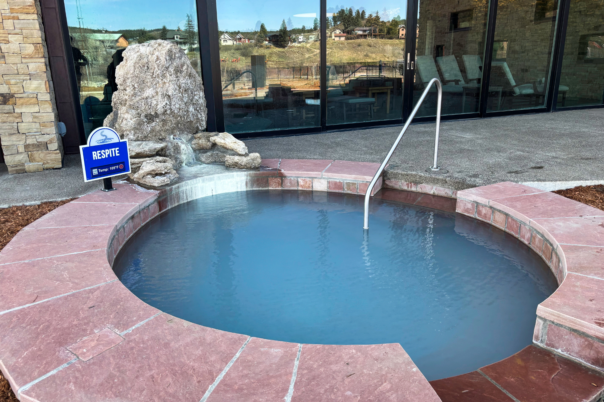 A circular outdoor hot tub contains milky blue water and a metal handrail, adjacent to a rock waterfall feature next to a modern building. Text: RESPITE, Temp: 105°F.