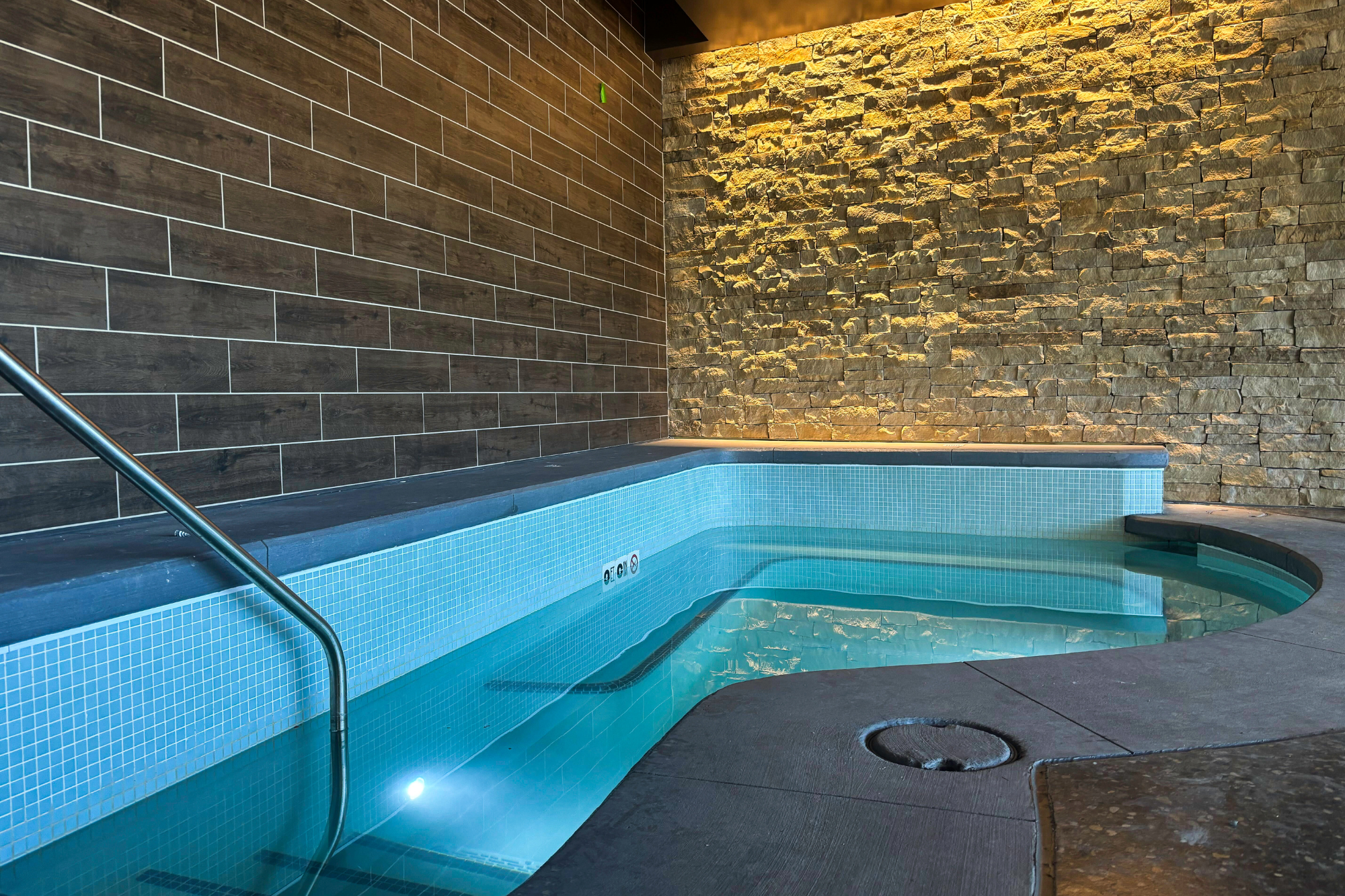 Indoor, curved pool with light blue mosaic tiles, clear water, and a metal handrail. Walls are dark wood-tiled and illuminated rough stone. '10' is on the pool wall.