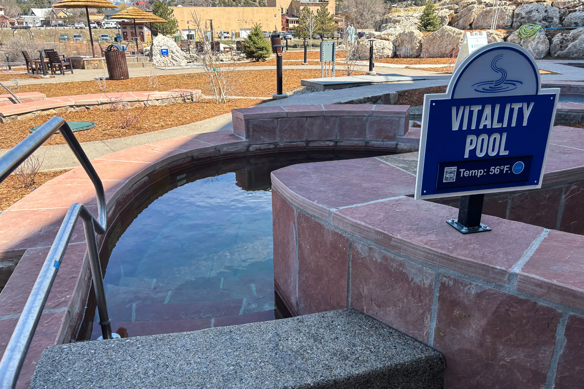 An outdoor stone pool holds clear water with steps and a handrail, within a landscaped public space featuring pathways and bare trees. Text on sign: VITALITY POOL, Temp: 56°F. (Cold), SCANNING TIPS.