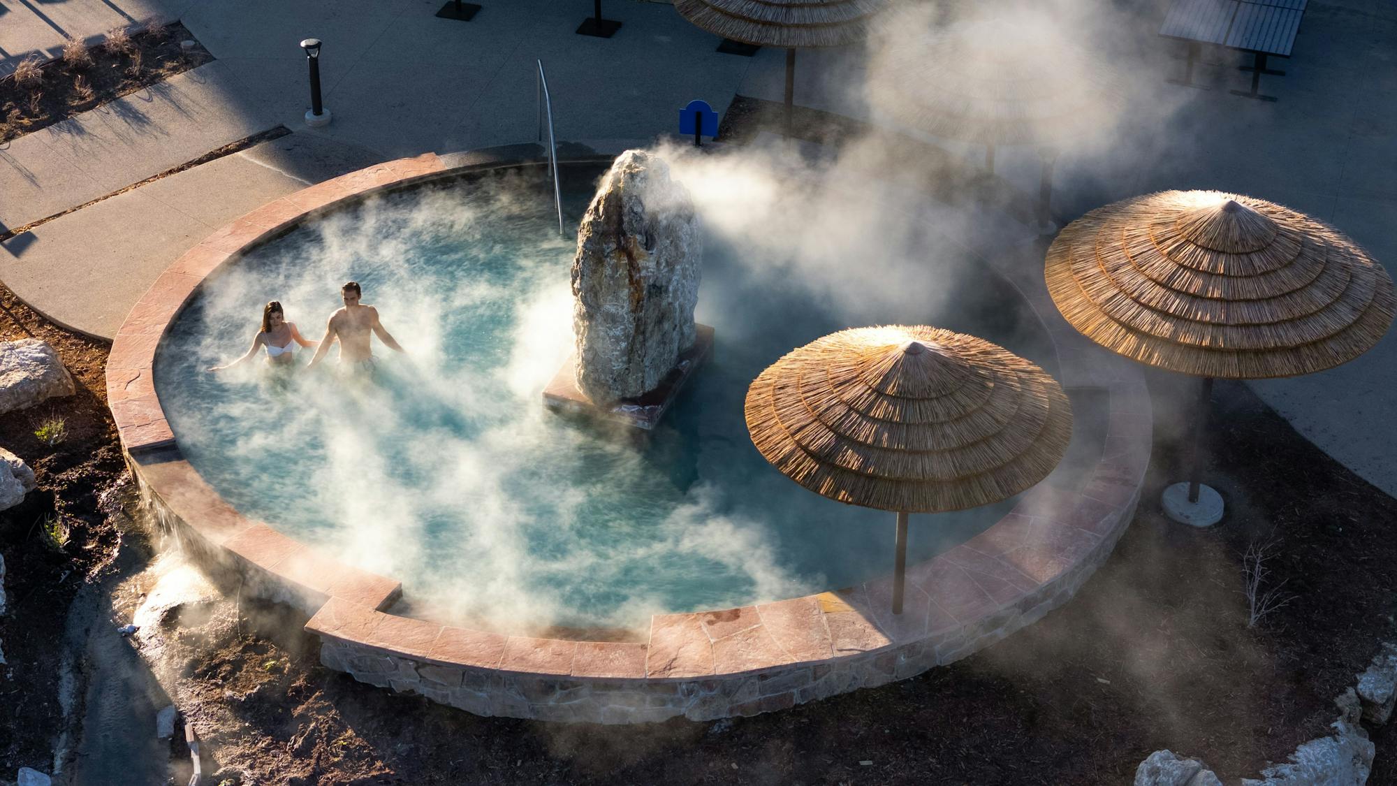 A couple relaxes in a steaming, circular outdoor hot spring with a large rock formation in the center. Two straw umbrellas stand nearby on concrete and natural ground.