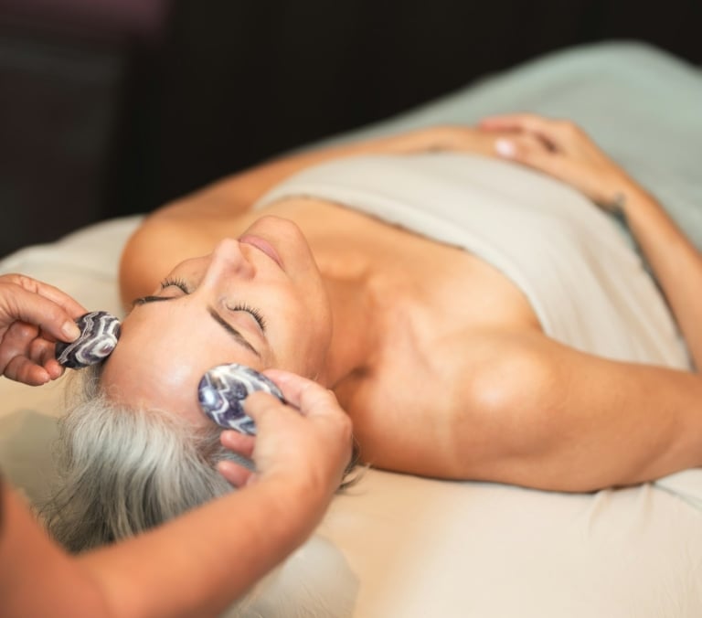 Woman with closed eyes receives a facial massage, as hands gently rub her forehead and temple with patterned purple and white stones on a massage bed.