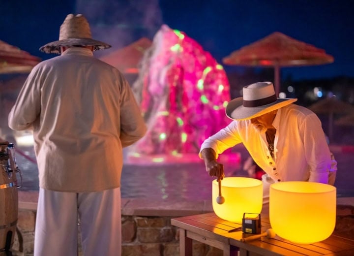 A man plays a glowing yellow singing bowl while another stands facing a brightly lit pink and green fountain at night, with beach umbrellas. A black box on the table reads: 