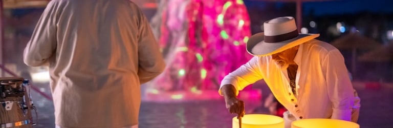A man plays a glowing yellow singing bowl while another stands facing a brightly lit pink and green fountain at night, with beach umbrellas. A black box on the table reads: 