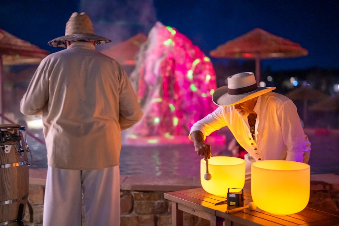 A man plays a glowing yellow singing bowl while another stands facing a brightly lit pink and green fountain at night, with beach umbrellas. A black box on the table reads: 