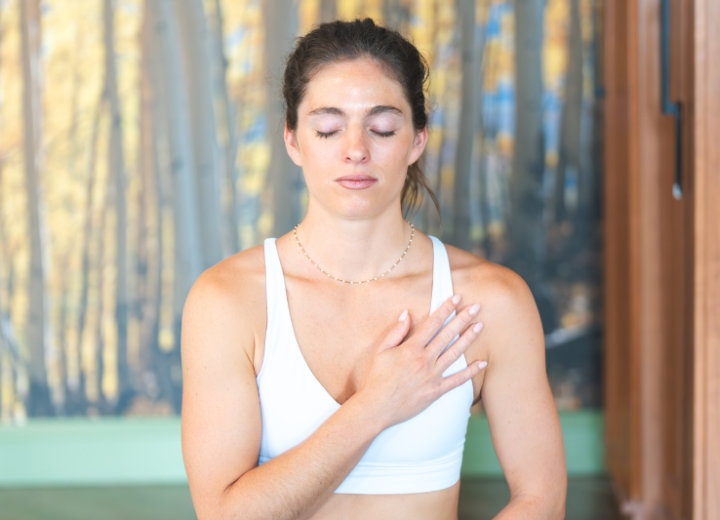 A young woman meditates with closed eyes and her right hand on her chest, wearing a white sports bra in front of a blurry aspen tree mural.