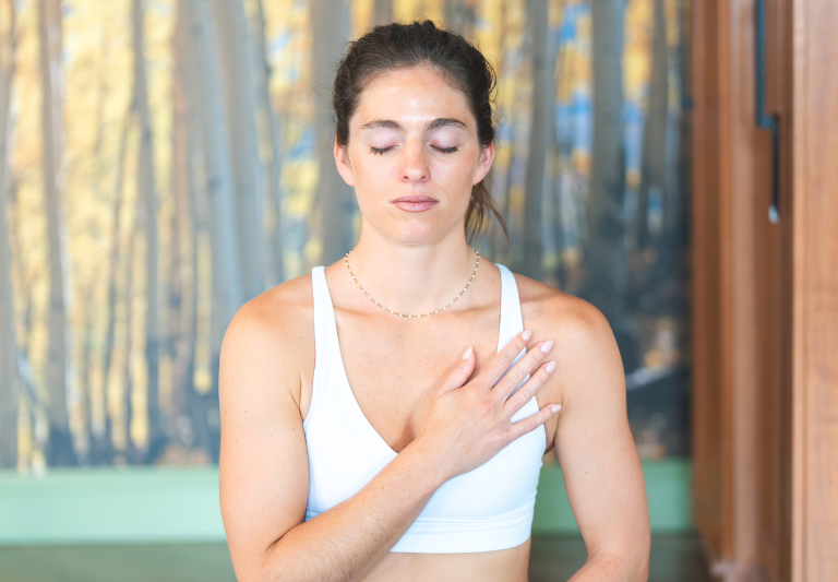 A young woman meditates with closed eyes and her right hand on her chest, wearing a white sports bra in front of a blurry aspen tree mural.