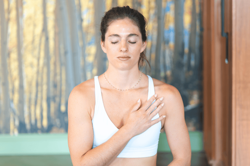 A young woman meditates with closed eyes and her right hand on her chest, wearing a white sports bra in front of a blurry aspen tree mural.