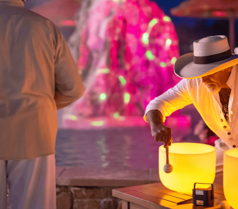 A man in a white hat and shirt strikes a glowing yellow crystal singing bowl. Another person stands nearby. They are outdoors at dusk, with a pink/green illuminated water feature in the background.
