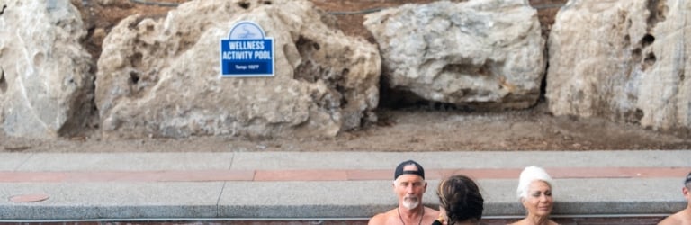 Three adults relax in a light blue activity pool, holding golden singing bowls, with large rock formations behind them. A sign reads: 