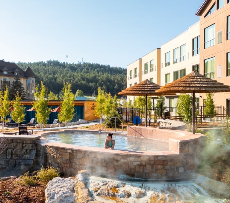A woman soaks in a steaming outdoor hot spring pool, near a building marked 