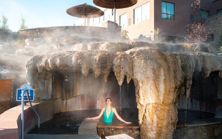 A woman in a green swimsuit relaxes in a steaming outdoor pool under a rock grotto. Straw umbrellas and a building are visible behind. Text: MEN'S GROTTO TEMP: 104F.