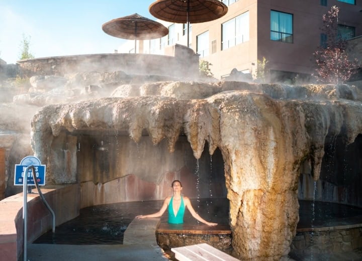 A woman in a green swimsuit relaxes in a steaming outdoor pool under a rock grotto. Straw umbrellas and a building are visible behind. Text: MEN'S GROTTO TEMP: 104F.
