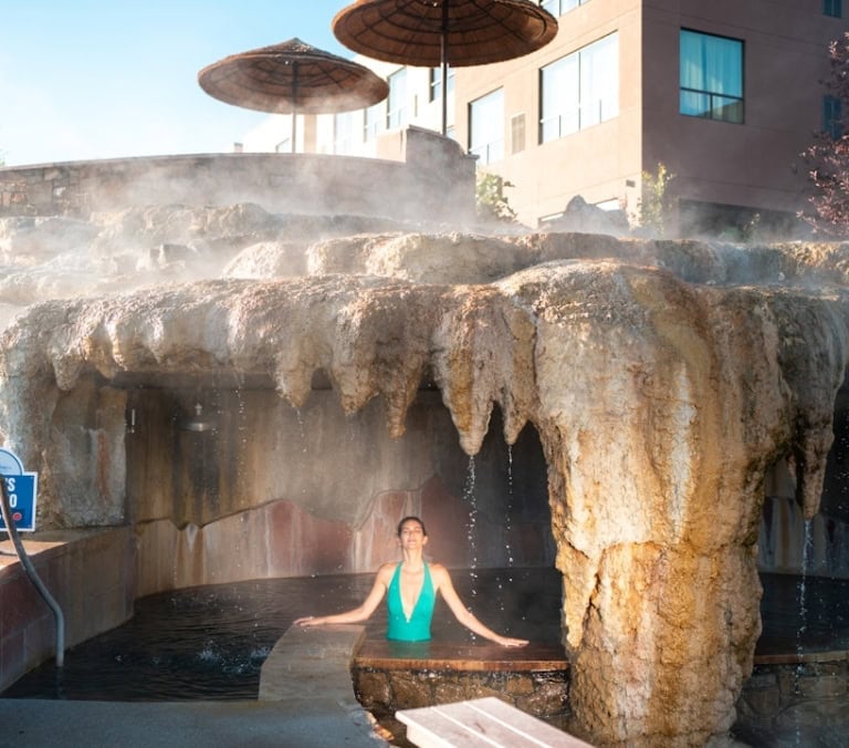 A woman in a green swimsuit relaxes in a steaming outdoor pool under a rock grotto. Straw umbrellas and a building are visible behind. Text: MEN'S GROTTO TEMP: 104F.