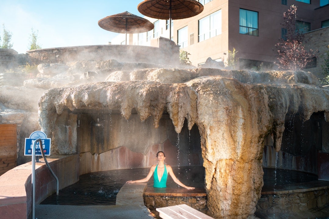 A woman in a green swimsuit relaxes in a steaming outdoor pool under a rock grotto. Straw umbrellas and a building are visible behind. Text: MEN'S GROTTO TEMP: 104F.