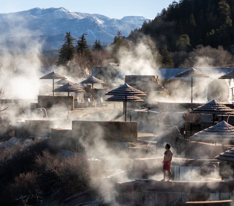 Man in red shorts stands amidst steaming hot spring pools with straw umbrellas, a river, and distant snow-capped mountains.