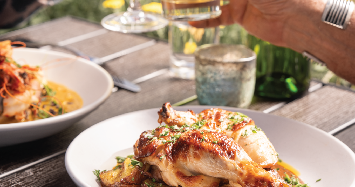 Roasted chicken and potatoes on a white plate, with a hand raising a beer glass in the background. Served on a wooden table with another shrimp dish, set outdoors.