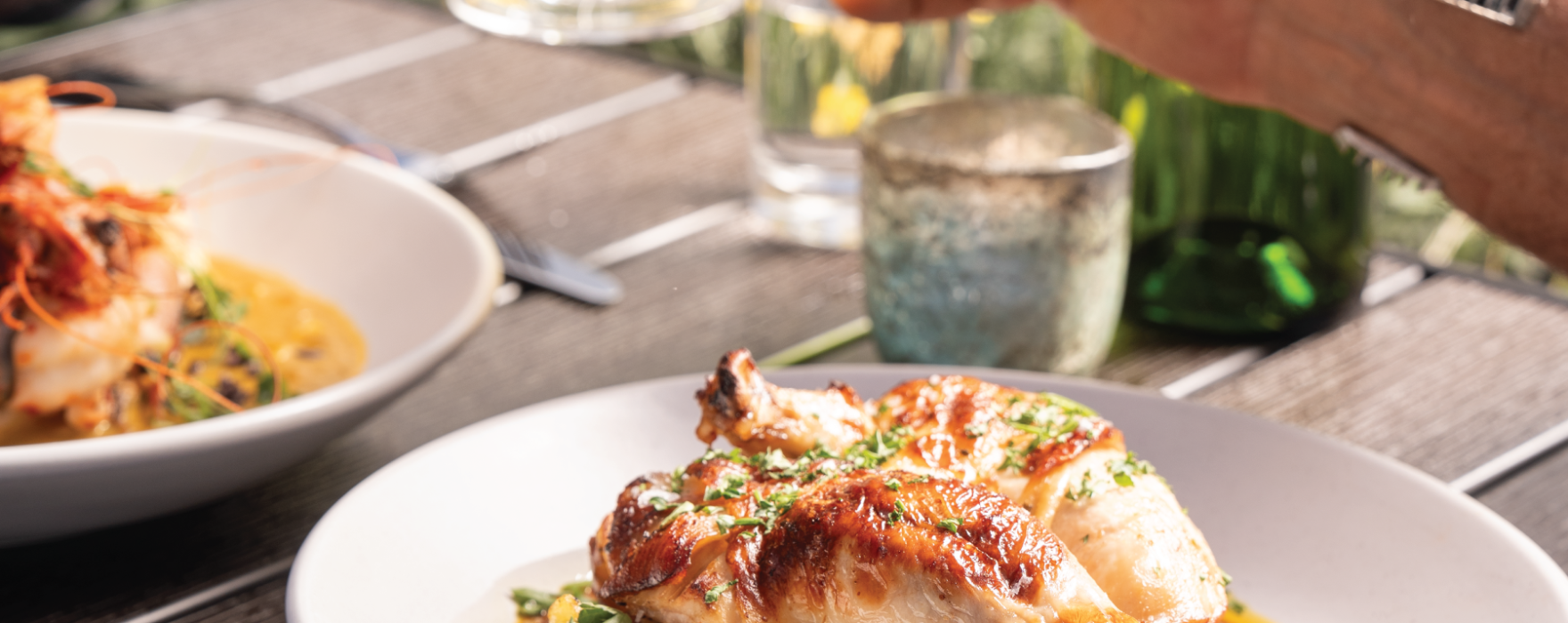 Roasted chicken and potatoes on a white plate, with a hand raising a beer glass in the background. Served on a wooden table with another shrimp dish, set outdoors.