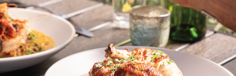 Roasted chicken and potatoes on a white plate, with a hand raising a beer glass in the background. Served on a wooden table with another shrimp dish, set outdoors.