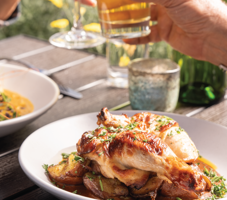 Roasted chicken and potatoes on a white plate, with a hand raising a beer glass in the background. Served on a wooden table with another shrimp dish, set outdoors.