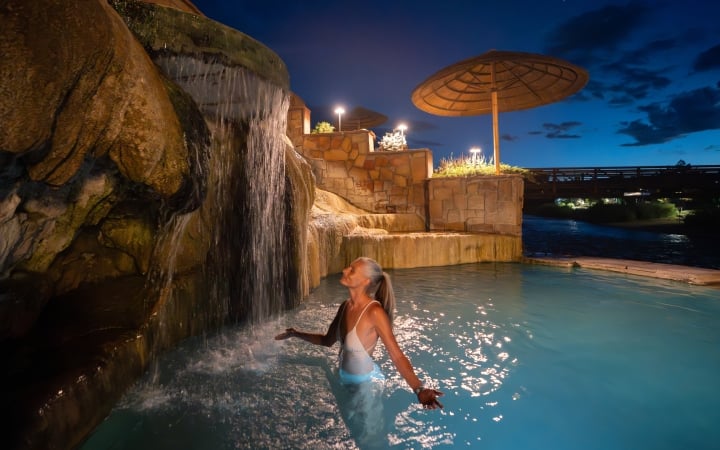 A woman with gray hair stands in a light blue pool, eyes closed, enjoying a rock waterfall. It's night, surrounded by illuminated stone walls and a distant bridge.