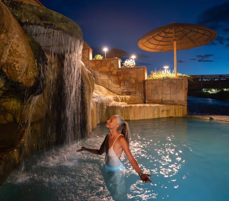 A woman with gray hair stands in a light blue pool, eyes closed, enjoying a rock waterfall. It's night, surrounded by illuminated stone walls and a distant bridge.