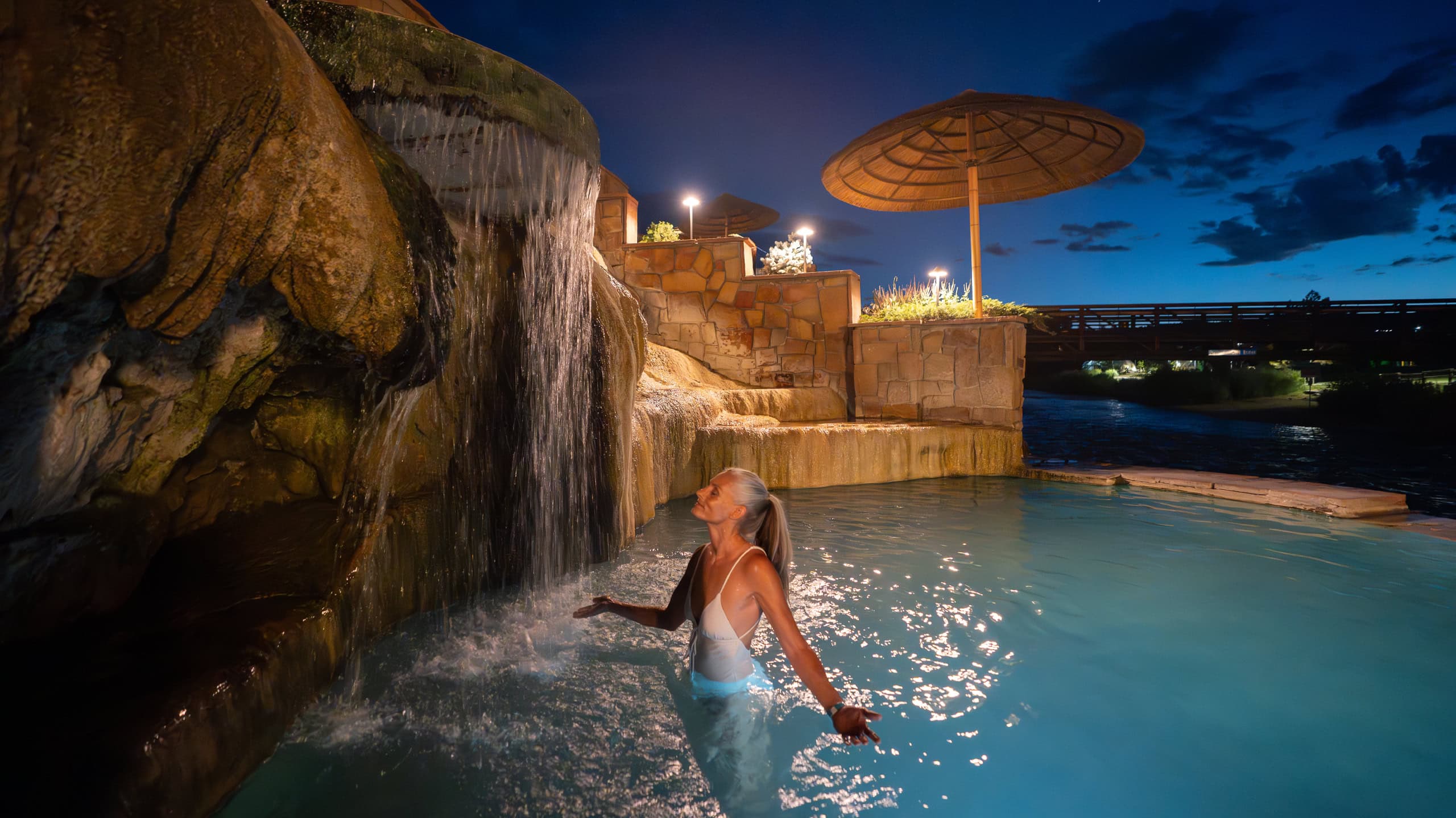 A woman with gray hair stands in a light blue pool, eyes closed, enjoying a rock waterfall. It's night, surrounded by illuminated stone walls and a distant bridge.