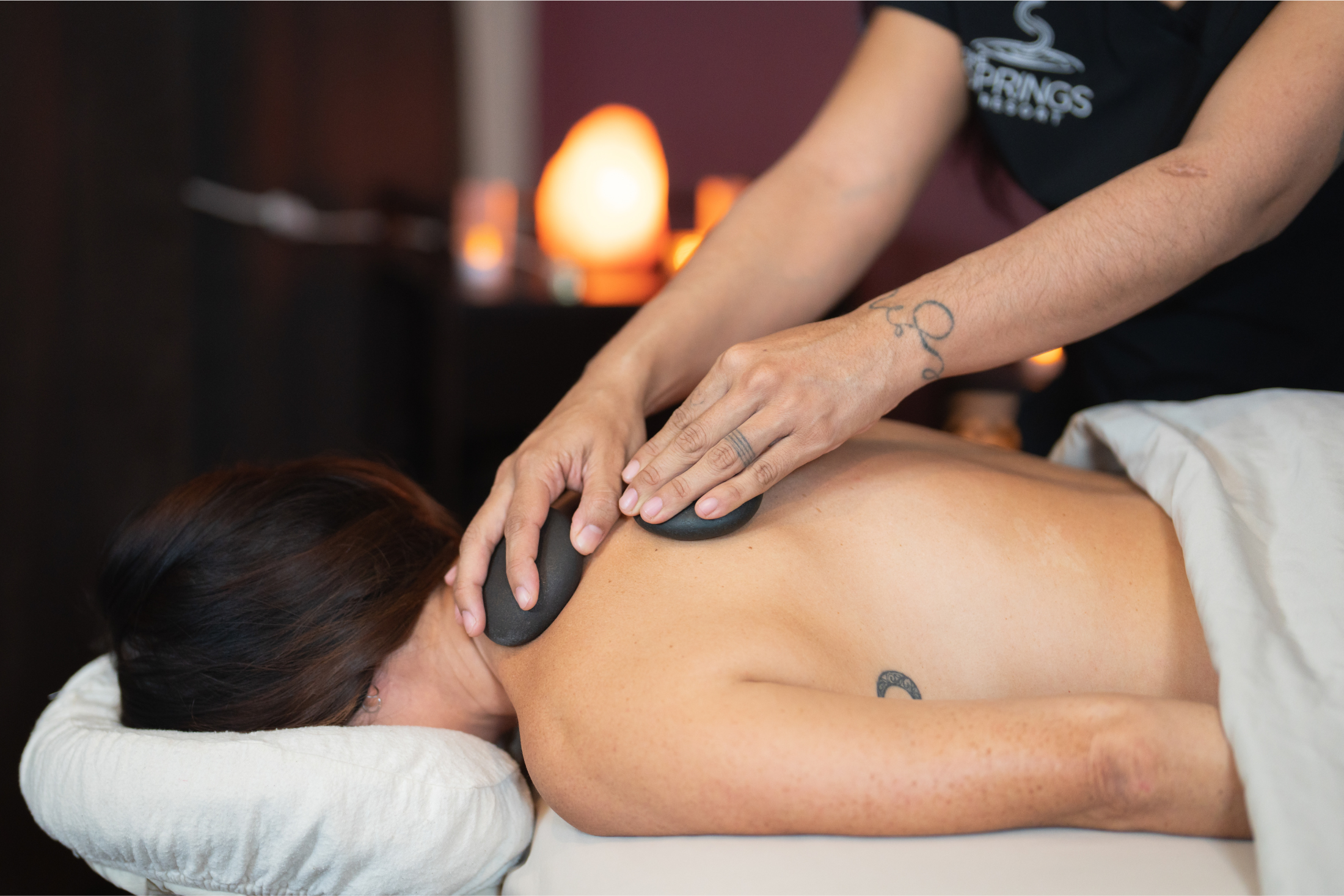 A person receives a hot stone massage as a masseuse places heated stones on their bare upper back in a dimly lit spa room. Masseuse's shirt: SPRINGS RESORT.