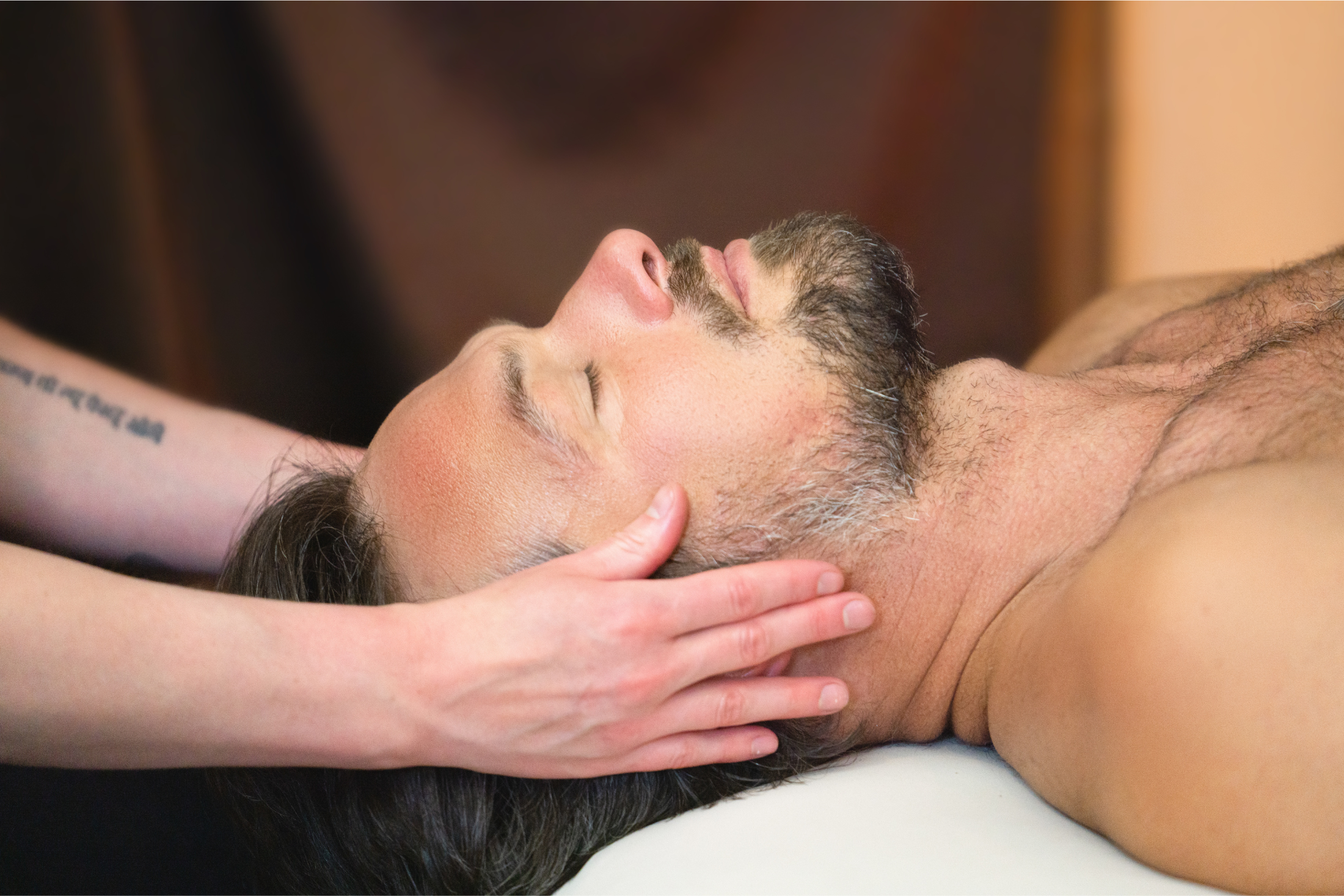 A bearded man with eyes closed receives a head massage from a person with a 