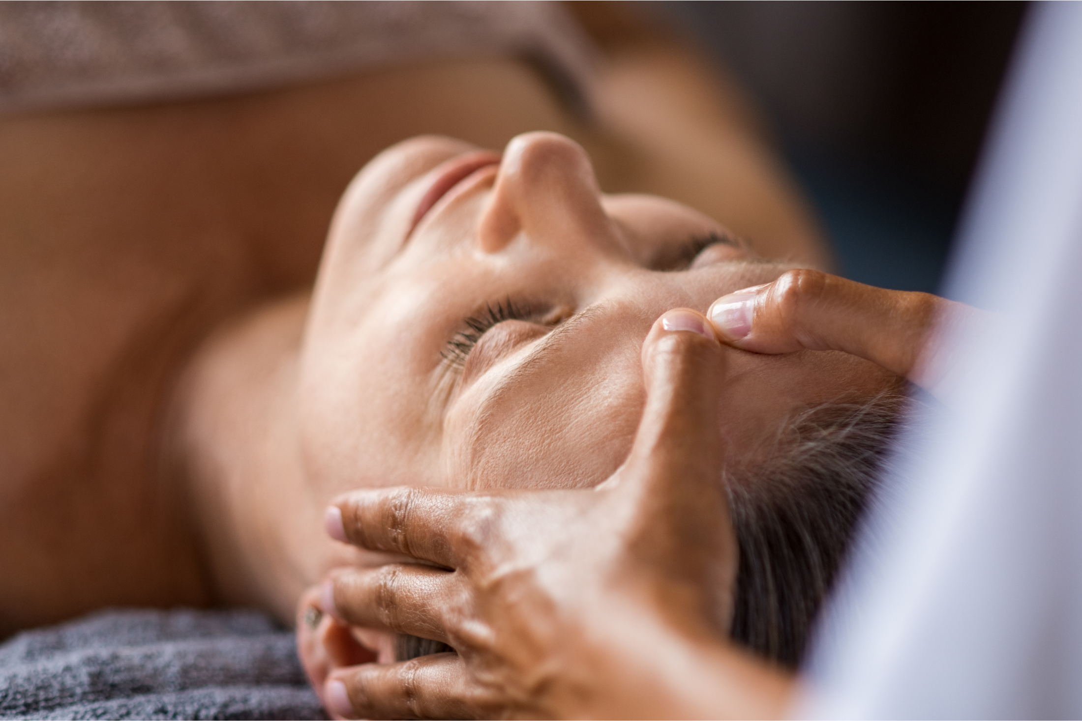 A woman with closed eyes receives a facial massage, with hands applying pressure to her temples, lying relaxed on a dark towel.