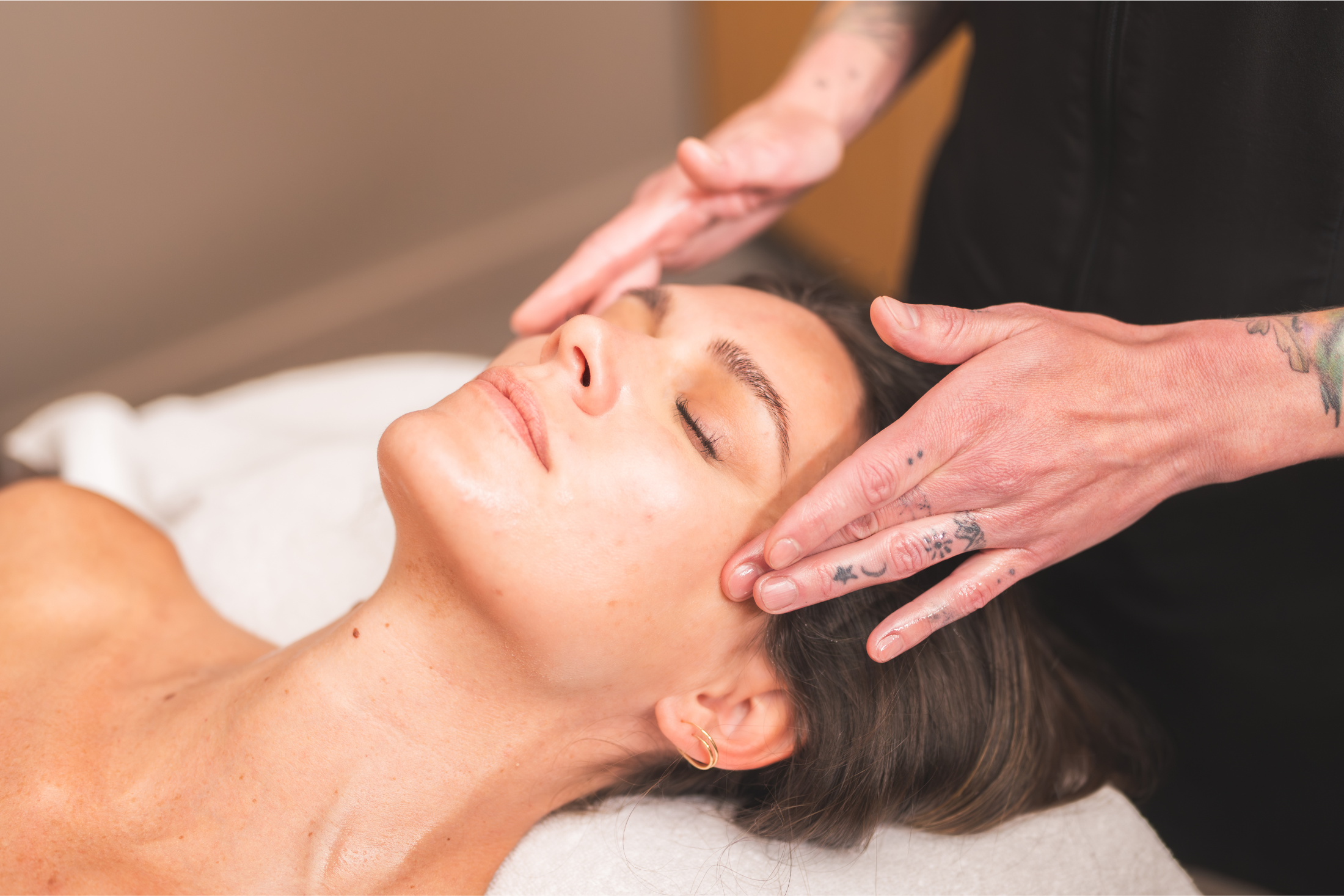 Woman with closed eyes receives a facial massage from tattooed hands, lying on a white towel in a soft-lit room.