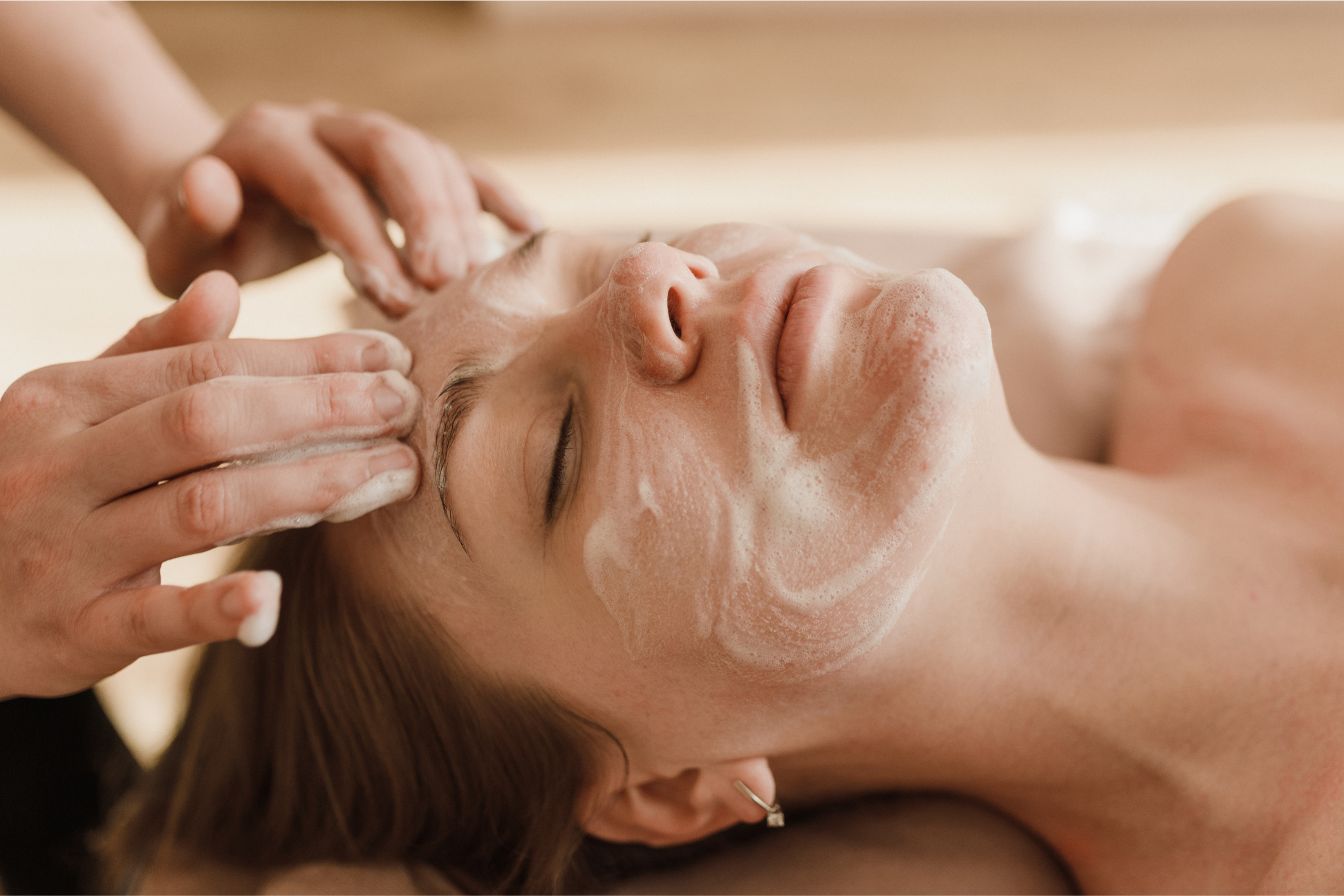 A woman lies relaxed with closed eyes, receiving a foamy facial treatment from two hands on her face in a bright, warm-toned spa.