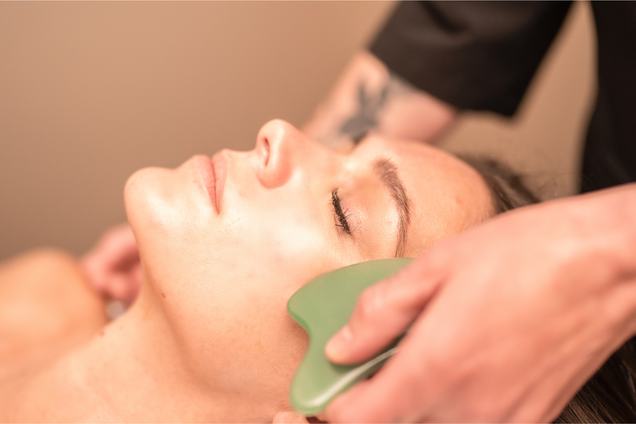 A woman lies with closed eyes, receiving a facial massage with a green Gua Sha tool applied by another's hand, in a soft-lit room.