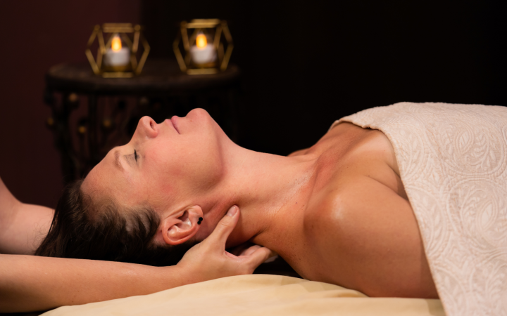 A person lies with closed eyes on a massage table, receiving a neck massage. A therapist's hands work on their neck. Two glowing candles illuminate a dark background.
