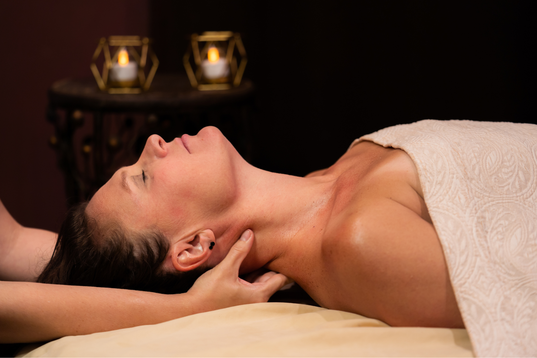 A person lies with closed eyes on a massage table, receiving a neck massage. A therapist's hands work on their neck. Two glowing candles illuminate a dark background.