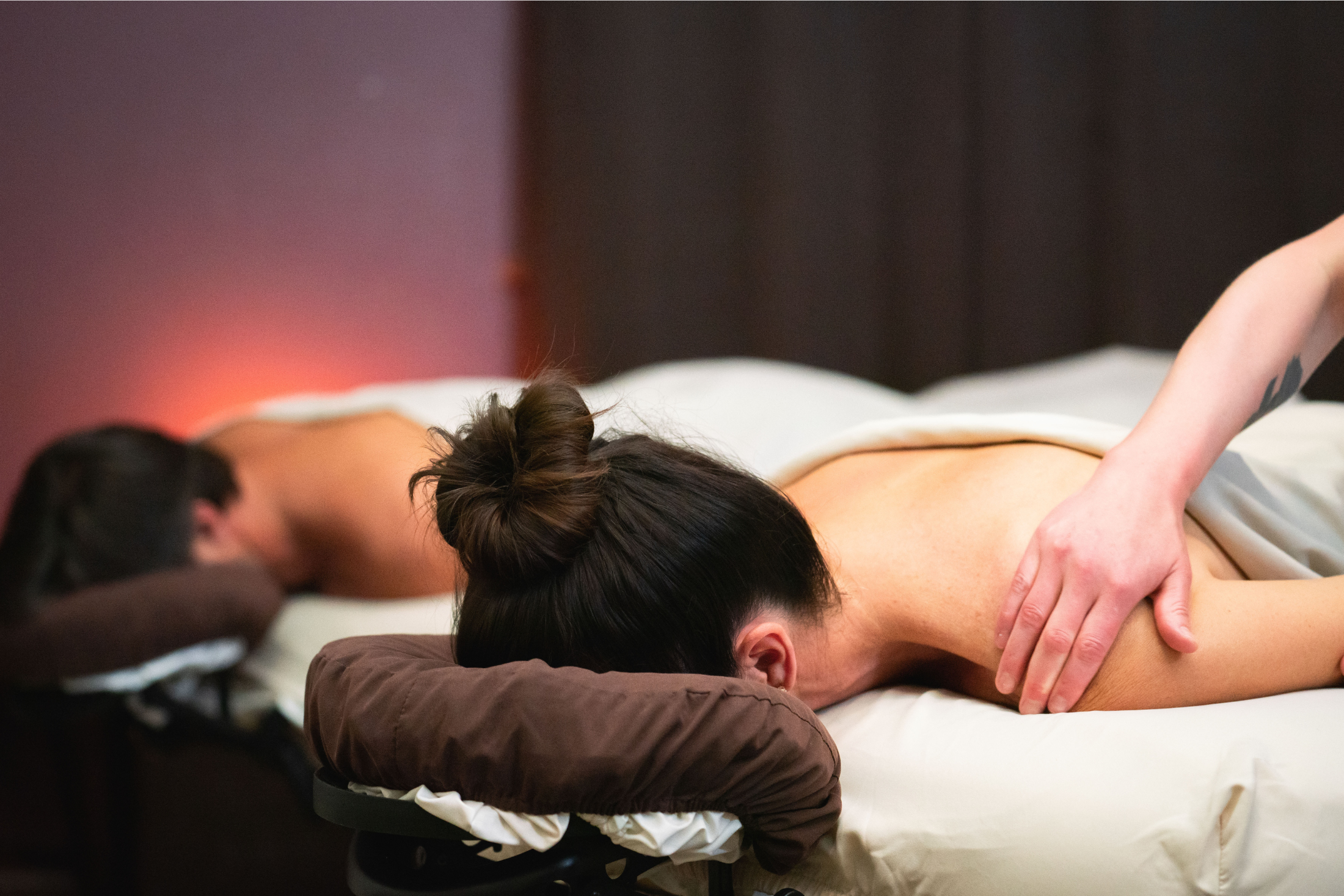 Two individuals receive massages, lying face down on separate beds in a dimly lit, warm-toned spa room. A hand massages the foreground person's shoulder.