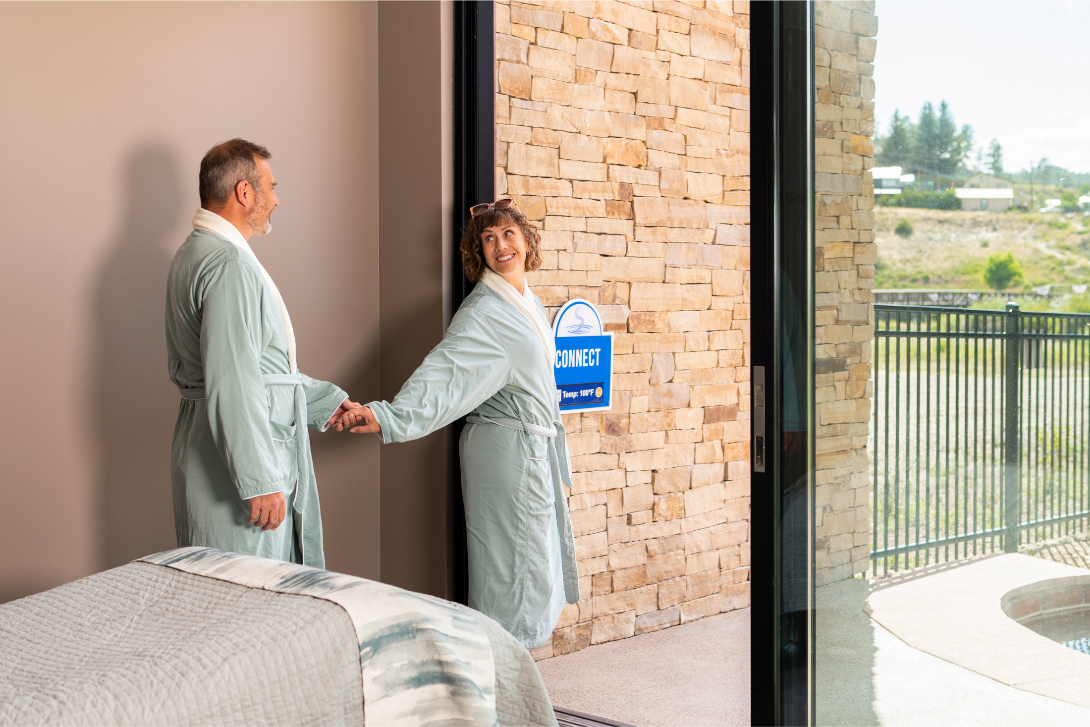 A couple in light green robes holds hands, stepping from a bedroom into an outdoor area with a stone wall and distant landscape. A blue sign reads: CONNECT Temp: 100°F.