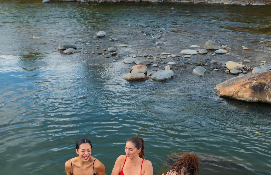 Three women laugh, standing chest-deep in a shallow river with visible rocks, framed by autumn trees with yellow and green foliage under a cloudy sky.