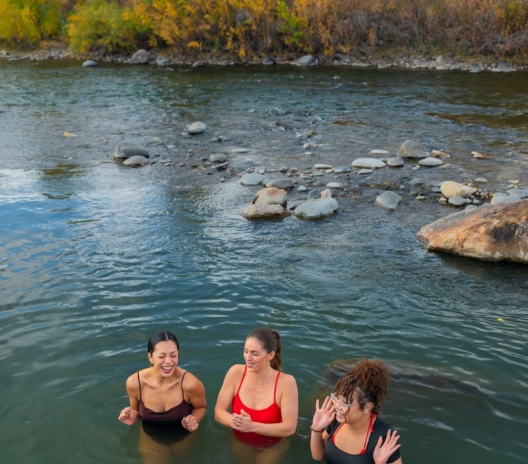Three women laugh, standing chest-deep in a shallow river with visible rocks, framed by autumn trees with yellow and green foliage under a cloudy sky.